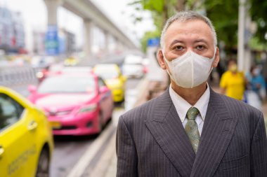 Portrait of mature Japanese businessman with mask for protection from corona virus outbreak at the taxi station in the city