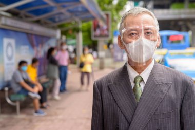 Portrait of mature Japanese businessman with mask and face shield for protection from corona virus outbreak at the bus stop