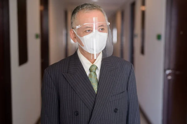 Portrait of mature Japanese businessman with mask and face shield for protection from corona virus outbreak in the corridor