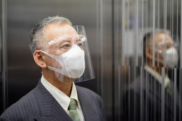 Portrait of mature Japanese businessman with mask and face shield for protection from corona virus outbreak at the elevator