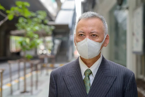 Portrait of mature Japanese businessman with mask for protection from corona virus outbreak in the city streets outdoors