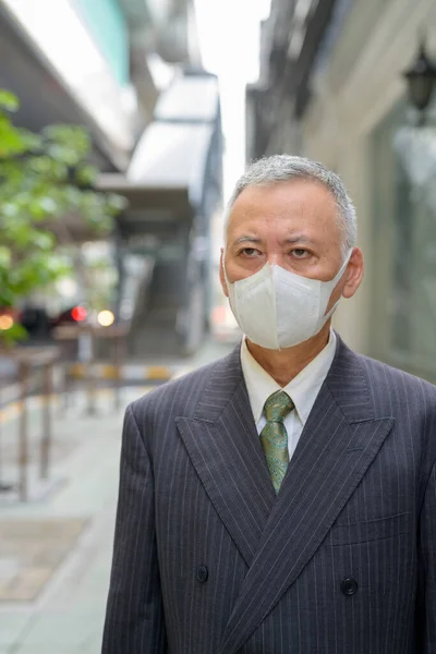 Portrait of mature Japanese businessman with mask for protection from corona virus outbreak in the city streets outdoors