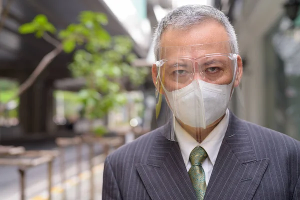 Portrait of mature Japanese businessman with mask and face shield for protection from corona virus outbreak in the city streets
