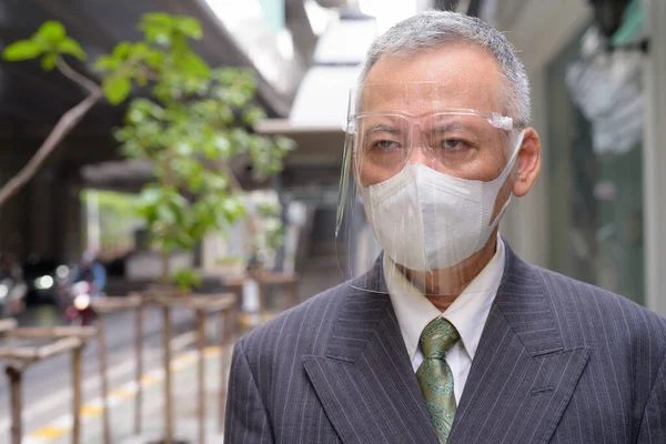 街中で発生したコロナウイルスから保護するためのマスクと顔の盾を持つ成熟した日本人ビジネスマンの肖像 — ストック写真