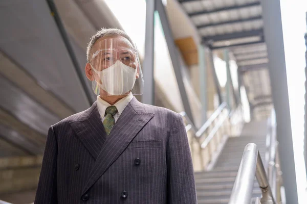 Portrait of mature Japanese businessman with mask and face shield for protection from corona virus outbreak in the city