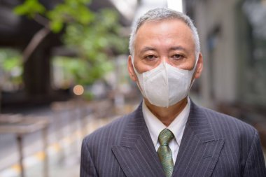 Portrait of mature Japanese businessman with mask for protection from corona virus outbreak in the city streets outdoors