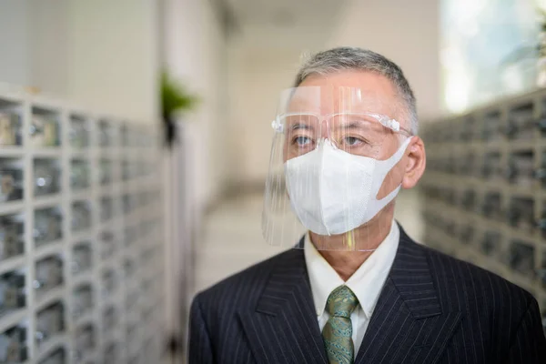 Portrait of mature Japanese businessman with mask and face shield for protection from corona virus outbreak at the mailbox