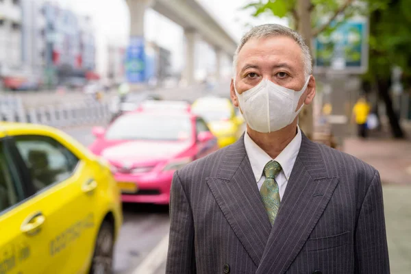 Portrait of mature Japanese businessman with mask for protection from corona virus outbreak at the taxi station in the city