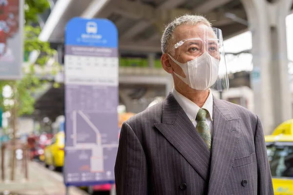 Portrait of mature Japanese businessman with mask and face shield for protection from corona virus outbreak at the bus stop