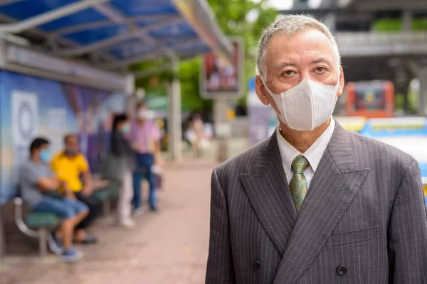 Portrait of mature Japanese businessman with mask for protection from corona virus outbreak at the bus stop in the city