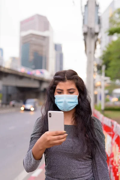 Portrait of young Indian woman with mask for protection from corona virus outbreak against view of the city streets outdoors