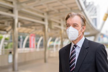 Portrait of mature businessman in suit with mask for protection from corona virus outbreak at skywalk bridge in the city outdoors