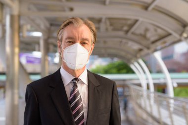 Portrait of mature businessman in suit with mask for protection from corona virus outbreak at skywalk bridge in the city outdoors