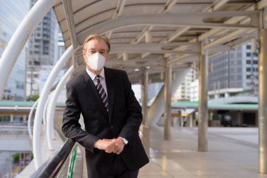 Portrait of mature businessman in suit with mask for protection from corona virus outbreak at skywalk bridge in the city outdoors