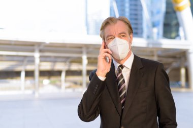 Portrait of mature businessman in suit with mask for protection from corona virus outbreak at skywalk bridge in the city outdoors