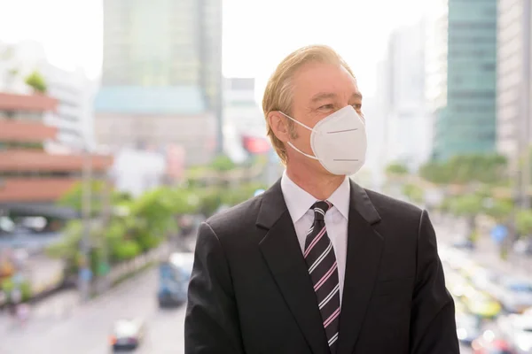 Portrait of mature businessman in suit with mask for protection from corona virus outbreak against view of the city