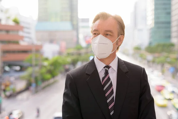 Portrait of mature businessman in suit with mask for protection from corona virus outbreak against view of the city