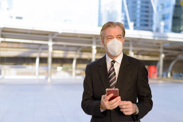Portrait of mature businessman in suit with mask for protection from corona virus outbreak at skywalk bridge in the city outdoors