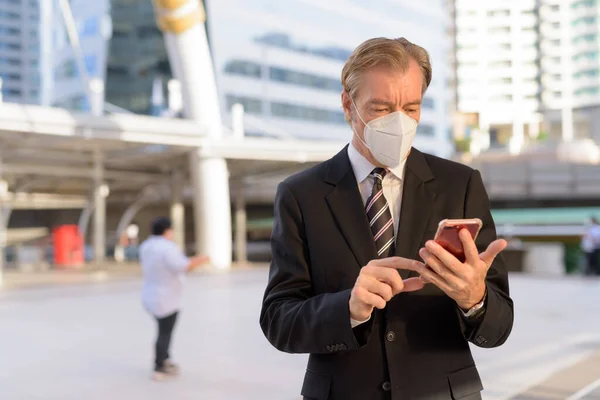 Portrait of mature businessman in suit with mask for protection from corona virus outbreak at skywalk bridge in the city outdoors