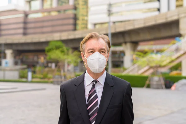 Portrait of mature businessman in suit with mask for protection from corona virus outbreak in the city streets outdoors