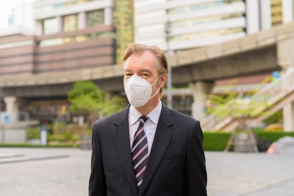 Portrait of mature businessman in suit with mask for protection from corona virus outbreak in the city streets outdoors