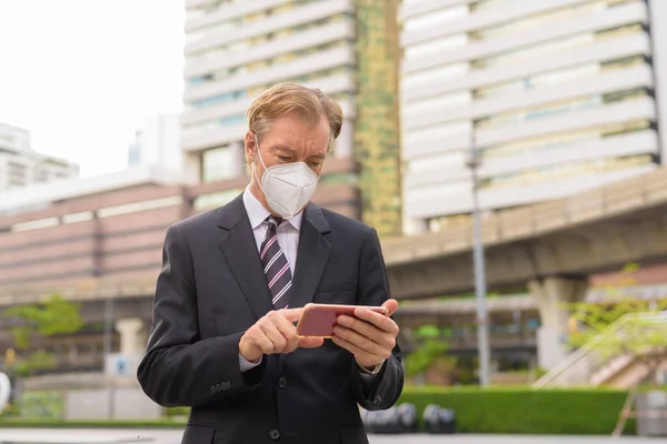 Érett üzletember maszkot használ telefon a városban a szabadban — Stock Fotó