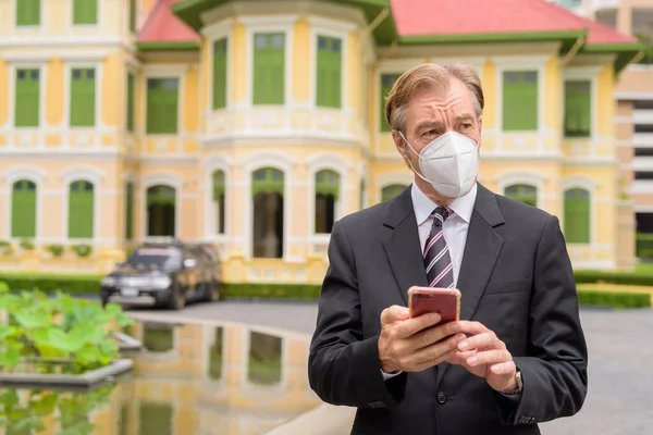 Homme d'affaires mature avec masque pensant tout en utilisant le téléphone en ville à l'extérieur — Photo
