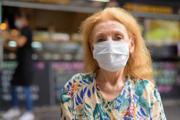 Szőke, idős nő maszkkal a koronavírus elleni védelemért a kávézóban. — Stock Fotó