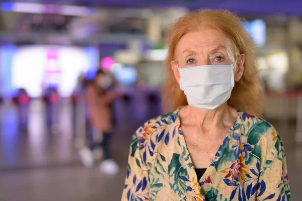 Egy szőke idős nő arca maszkban és társadalmi távolságban a vasútállomáson. — Stock Fotó