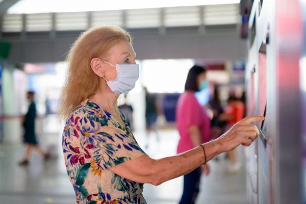 Blonda femeie în vârstă cu mască cumpărarea bilet la gara cer — Fotografie, imagine de stoc