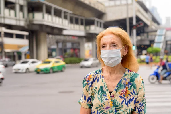 Blonde senior woman thinking and wearing mask for protection from corona virus outbreak in the city outdoors