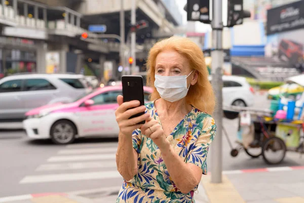 Szőke idős nő maszkot használ telefon a városban a szabadban — Stock Fotó