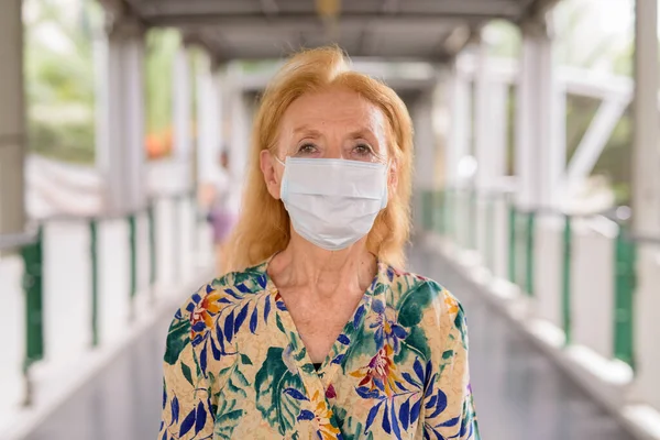 Egy szőke idős nő portréja maszkban a corona vírus kitörése elleni védelemért a hídnál. — Stock Fotó