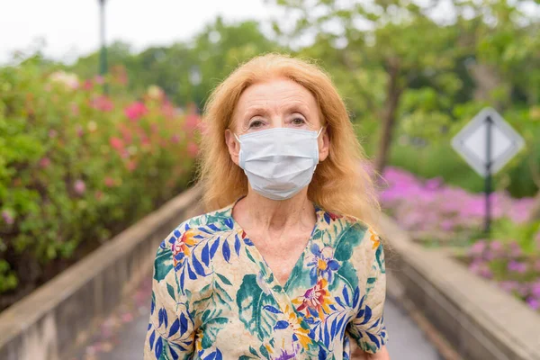 Egy szőke idős nő arca, aki maszkot visel, hogy megvédje magát a koronavírus kitörésétől a parkban. — Stock Fotó