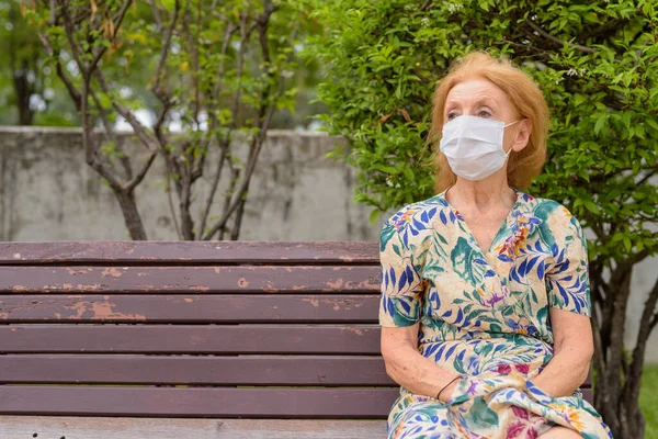 Portret de blonda senior femeie cu masca gândire în timp ce stând cu distanță pe banca de parc — Fotografie, imagine de stoc