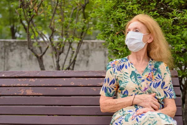 Portret de blonda senior femeie cu masca gândire în timp ce stând cu distanță pe banca de parc — Fotografie, imagine de stoc