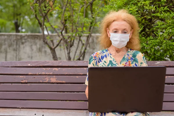 Portré szőke idős nő maszk segítségével laptop, miközben ül a távolság park padon — Stock Fotó