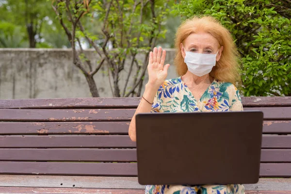 Szőke idős nő maszk videó hívás segítségével laptop, miközben ül a távolság park padon — Stock Fotó