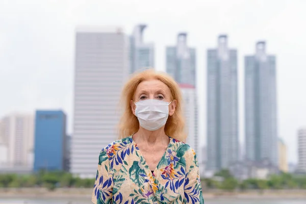 Egy szőke idős nő portréja, aki maszkot visel, hogy védve legyen a koronavírus kitörése ellen, és ne lássa a várost. — Stock Fotó