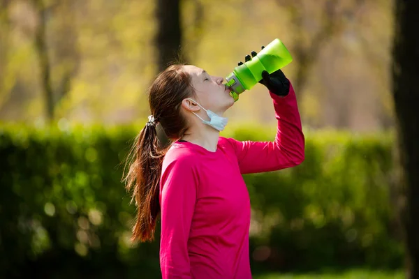 公園でスポーツをした後若い女の子は水を飲む 隔離後の医療用マスクと手袋の少女 ロイヤリティフリーのストック画像