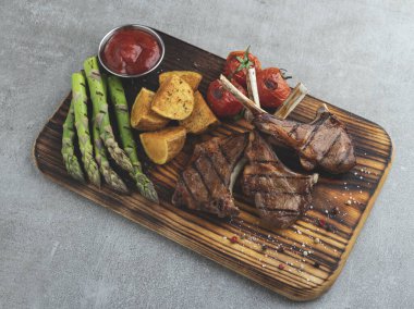 Ahşap bir tahtada ızgara kuzu pirzola, kuşkonmaz, kırmızı sos, patates, domates.