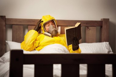 Adam yatakta yalnız başına, tehlikeli madde giysisi içinde kitap okurken maske takıyor. 