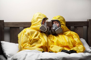 İki kişi yatakta yalnızlık çekiyor. Çok dikkatli davranıyorlar ve tehlikeli madde kıyafeti giyiyorlar.