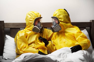 İki kişi yatakta yalnızlık çekiyor. Çok dikkatli davranıyorlar ve tehlikeli madde kıyafeti giyiyorlar.