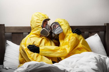 İki kişi yatakta yalnızlık çekiyor. Çok dikkatli davranıyorlar ve tehlikeli madde kıyafeti giyiyorlar.