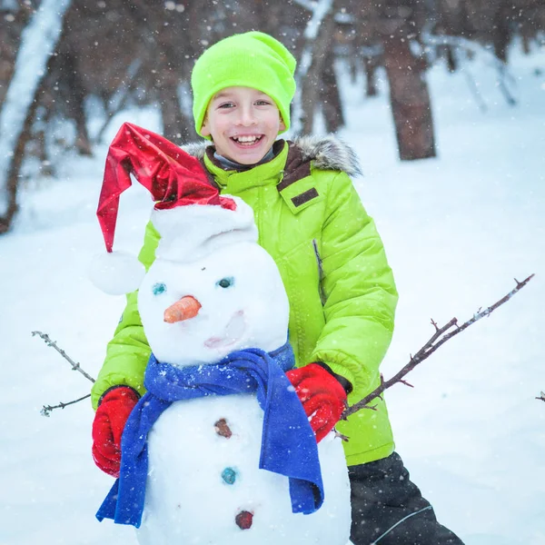 Веселый, счастливый малыш играет со снеговиком в зимнем парке — стоковое фото