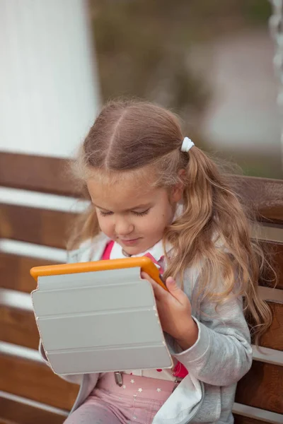 Ciddi bir kız tabletle meşgul, evin avlusundaki tahta bir bankta oturuyor. — Stok fotoğraf