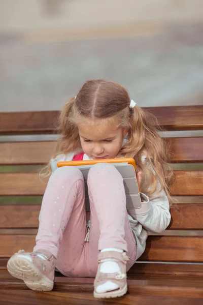 Chica seria se dedica a una tableta, ella está sentada en un banco de madera en el patio de la casa —  Fotos de Stock