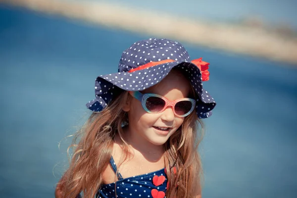 Портрет милой веселой, счастливой девушки в шляпе и солнцезащитных очках, на открытом воздухе. Девушка сидит на берегу моря . — стоковое фото