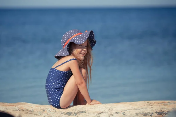 Açık havada şapka ve güneş gözlüğü takan sevimli, neşeli bir kızın portresi. Deniz kıyısında oturan kız. — Stok fotoğraf